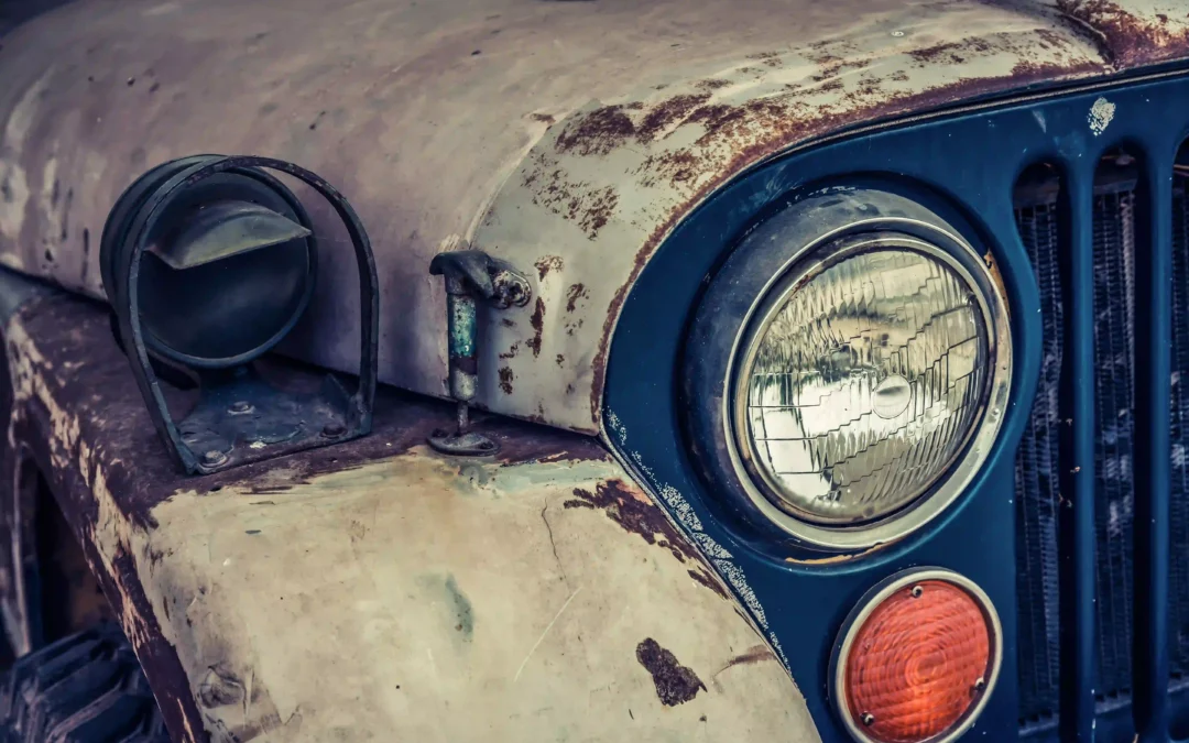 Rusty vintage car front close-up, with weathered paint, round headlight, and an aged orange indicator.