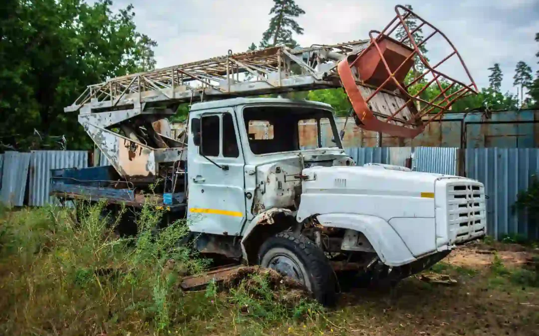 Abandoned white utility truck with rusted crane/basket lift, parked in overgrown grass beside a metal fence