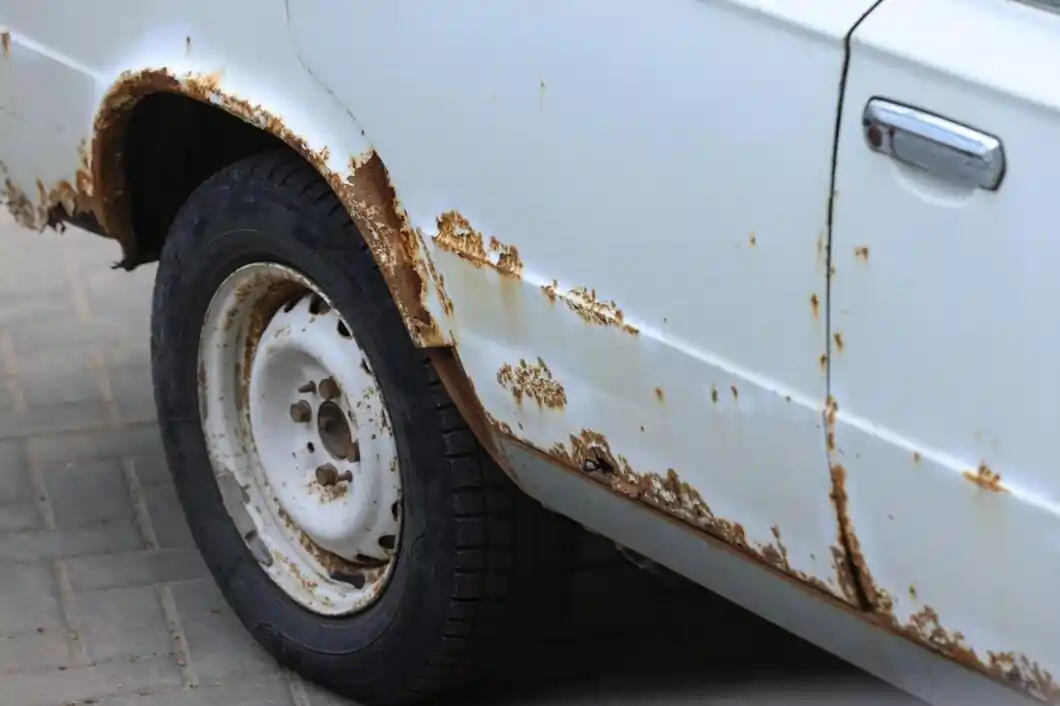 A white car's rocker panel and rear wheel arch showing heavy rust bubbling and paint peeling from road salt corrosion