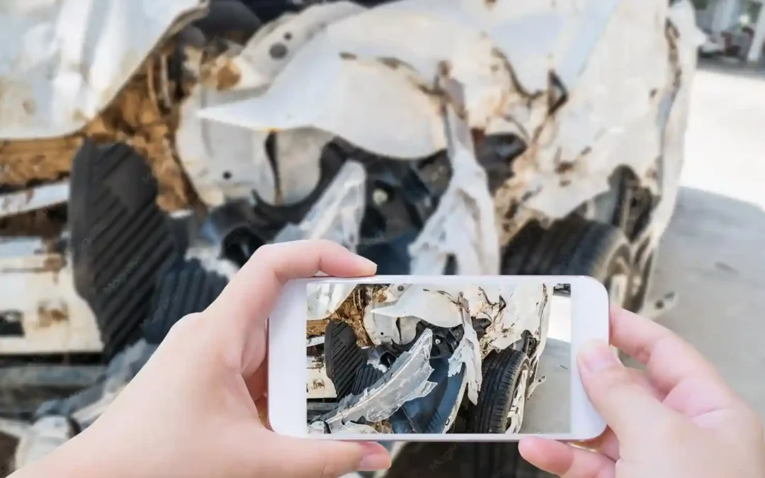 Hands hold a smartphone, photographing a wrecked car's crushed, rusted front end before scrapping it.