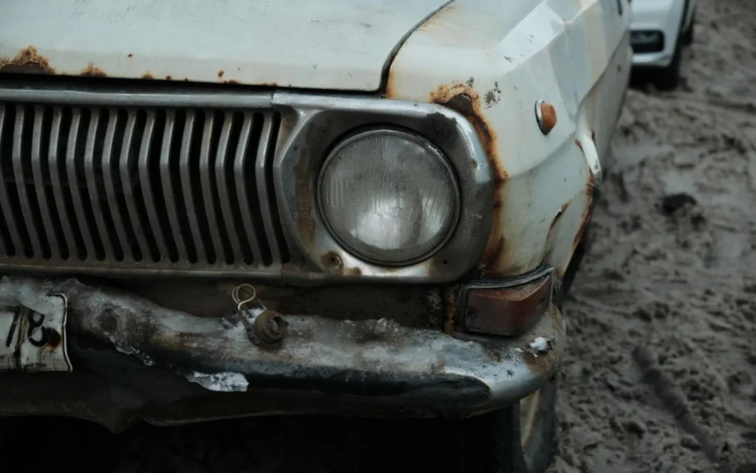 Close-up of a rusted, weathered car's front bumper and headlight, parked on muddy ground outdoors